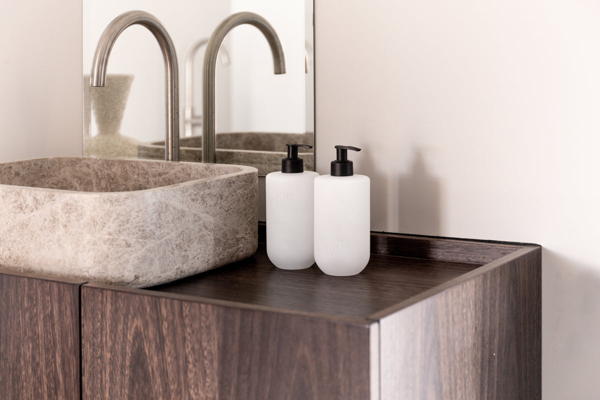 Landscape crop of frosted bottles on a dark timber vanity beside a rough stone basin