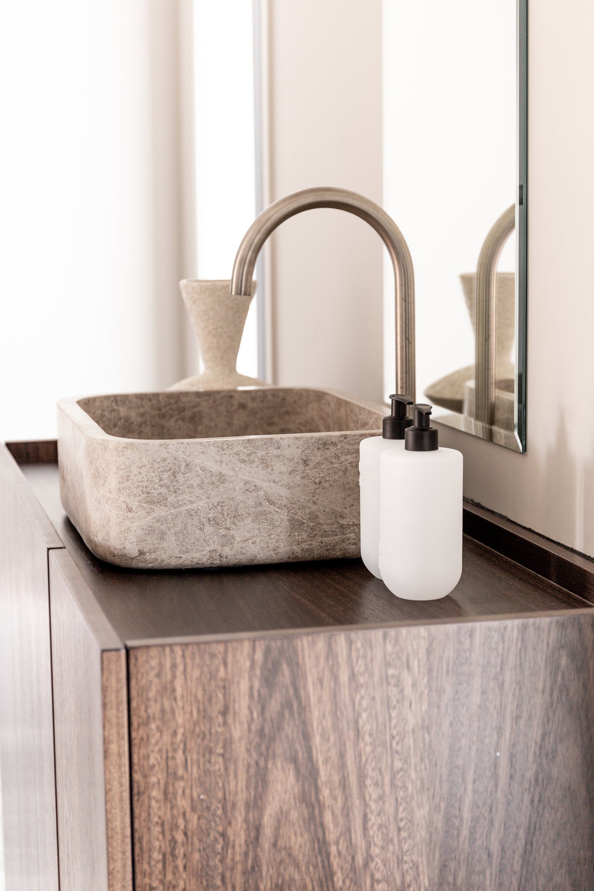 Frosted pump bottles beside a stone vessel basin and arched brass tap on a dark timber vanity