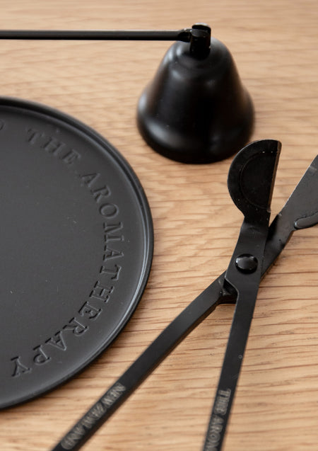 Black candle snuffer and wick trimmer beside an embossed black Therapy plate on light oak