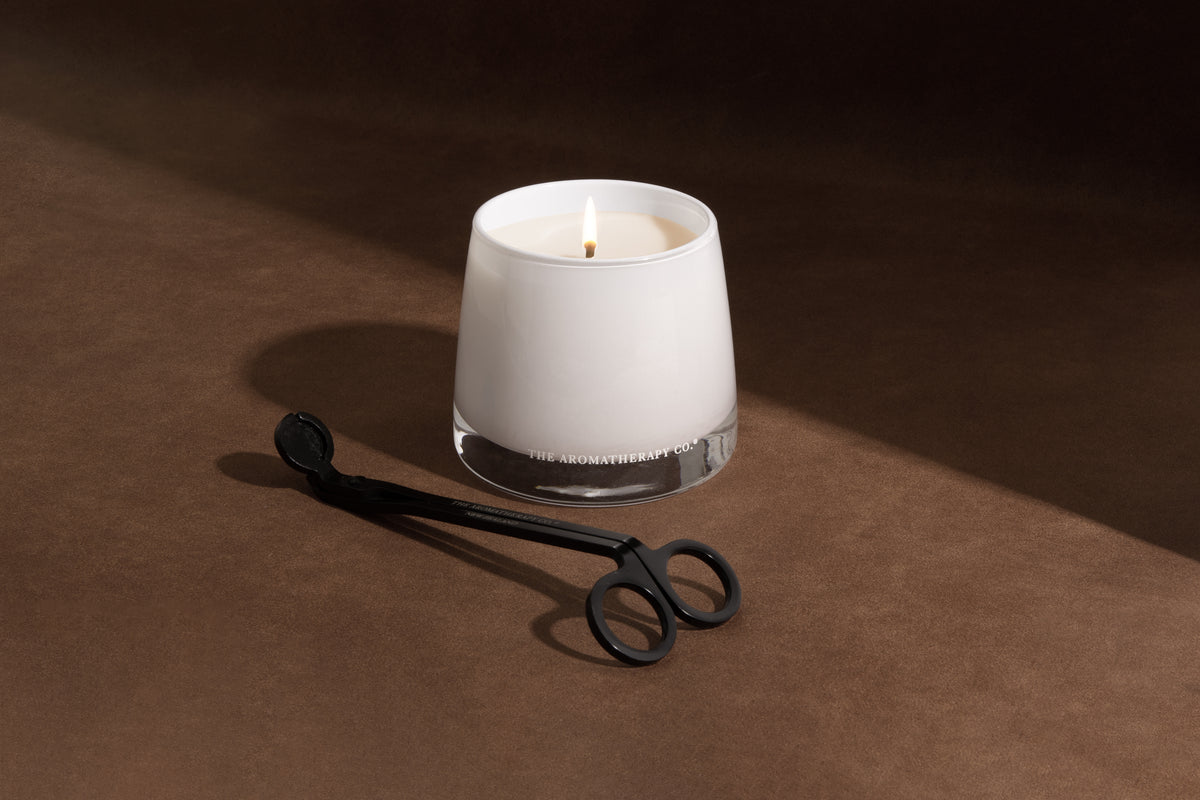 Lit Therapy candle in frosted white glass with a black wick trimmer resting in front on warm brown