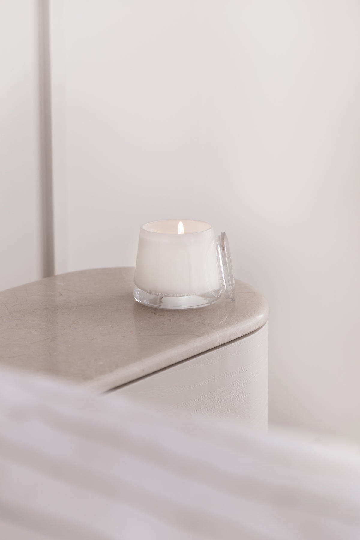 Lit Therapy candle on the corner of a pale curved side table beside a soft white curtain