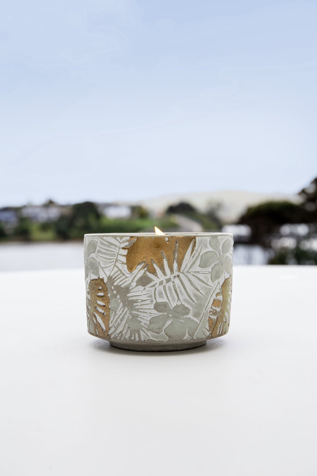 Leaf-embossed candle on a white table against a blue sky, with water and houses on the horizon