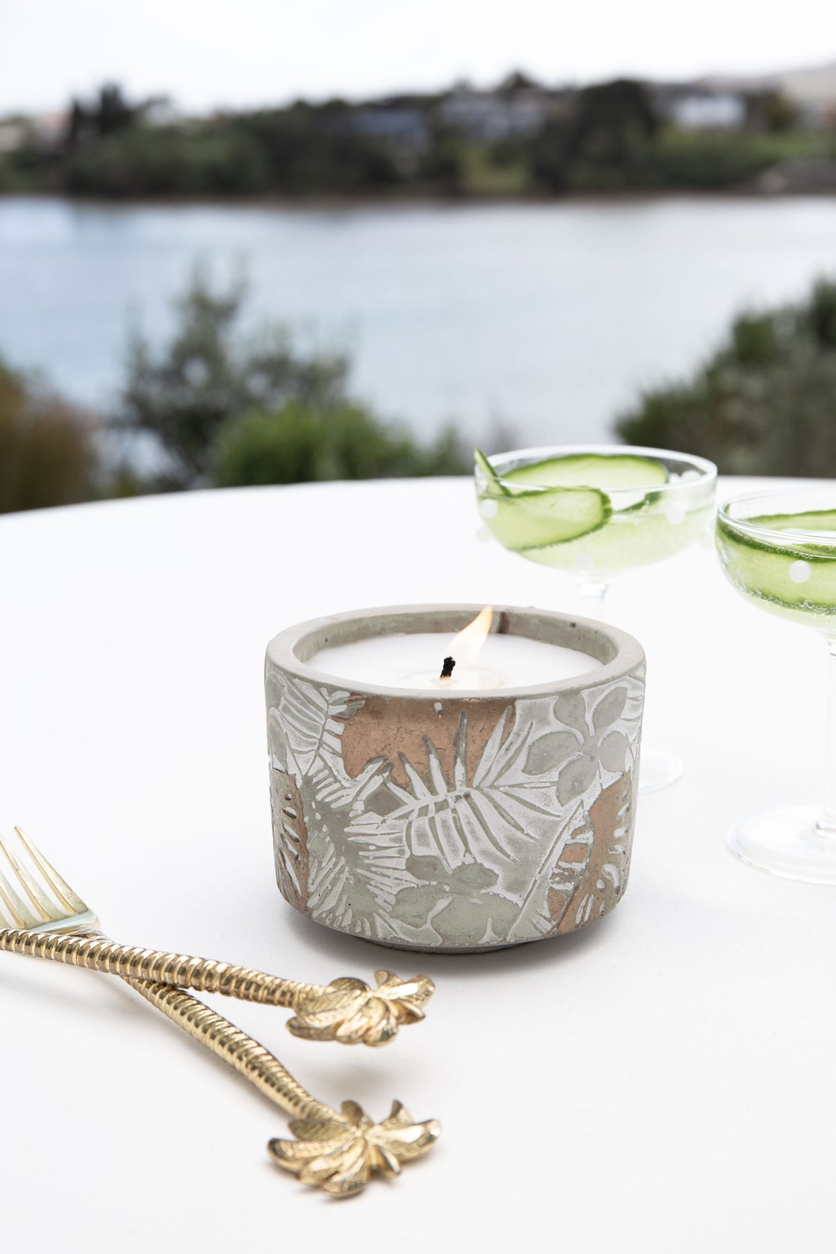 Lit candle on a white table by a lake with cucumber cocktails and gold palm forks