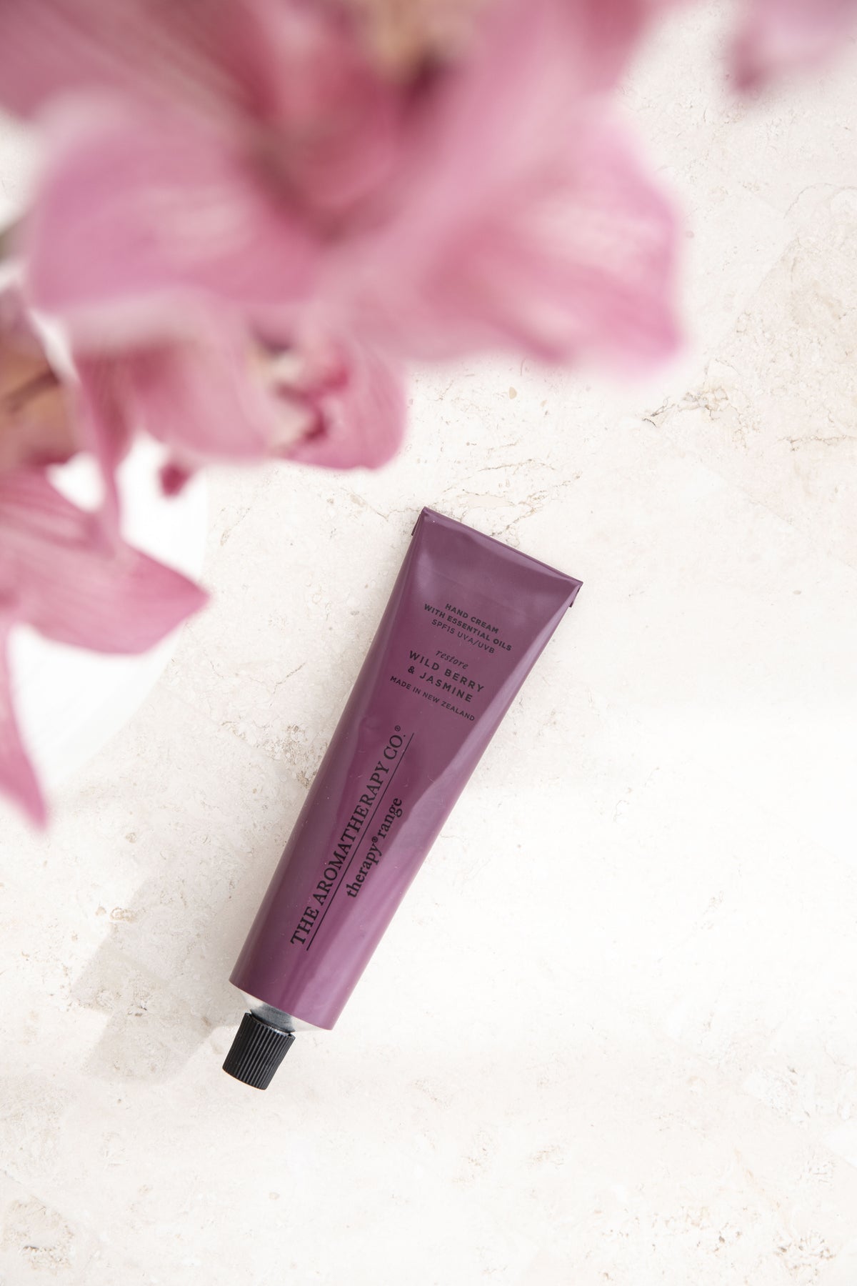 Plum hand cream tube lying diagonally on pale stone under soft focus pink orchids