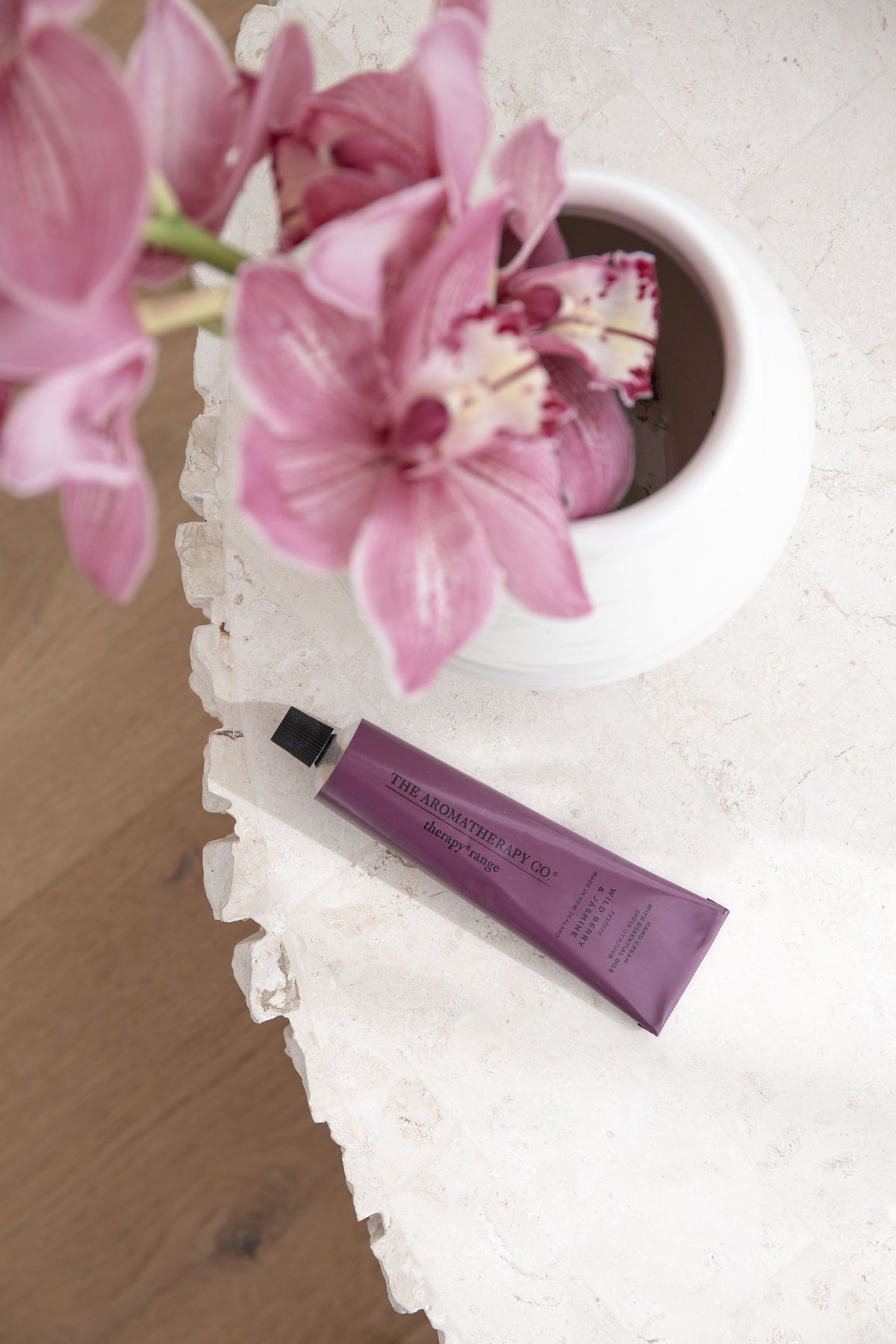 Plum hand cream tube on a stone plinth beside a white bowl of pink orchids
