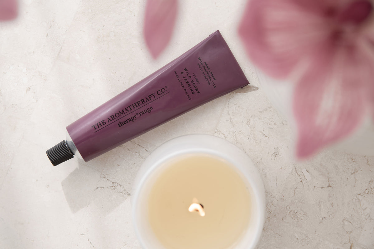 Landscape overhead of the hand cream tube, a lit candle and pink orchids on marble