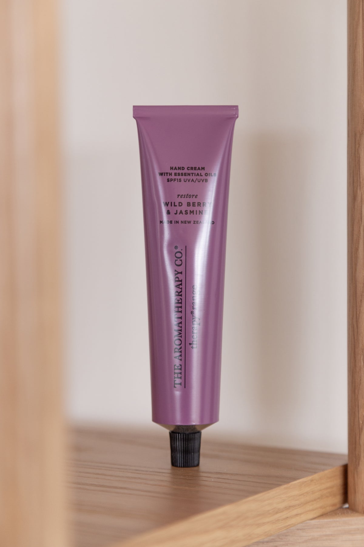 Plum hand cream tube standing in the corner of an oak shelf against a pale wall