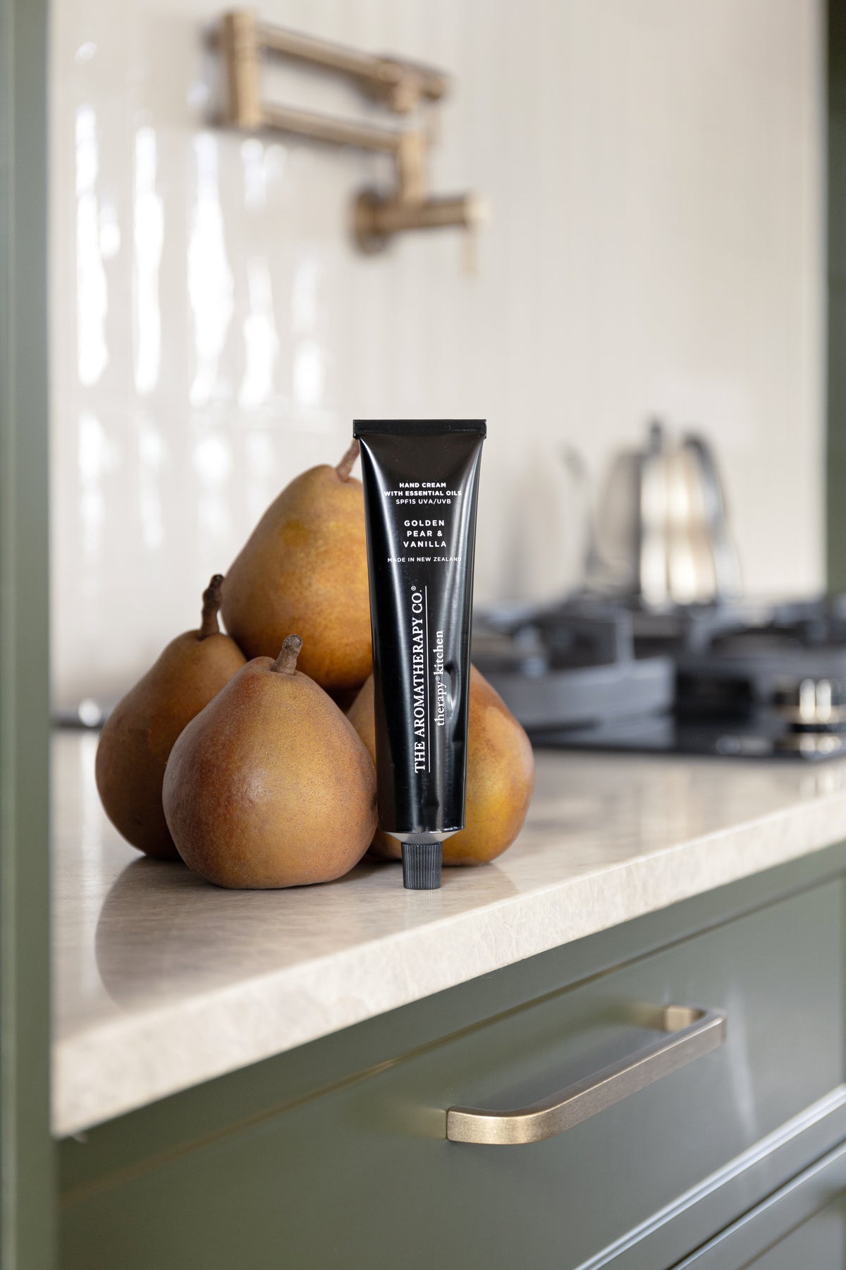 Hand cream tube beside a cluster of pears on a kitchen bench above an olive green drawer
