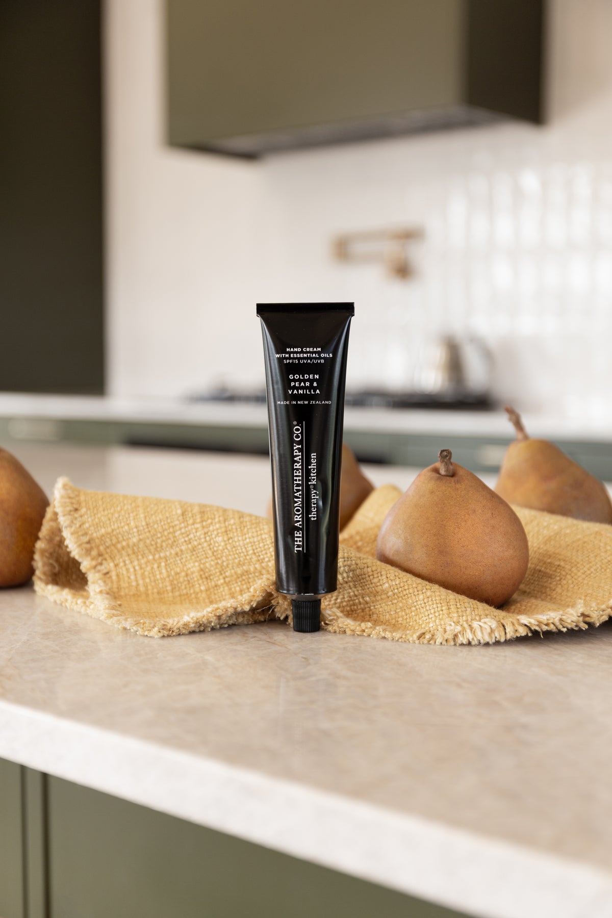 Hand cream tube on a stone bench beside pears resting on a mustard linen cloth, rangehood behind