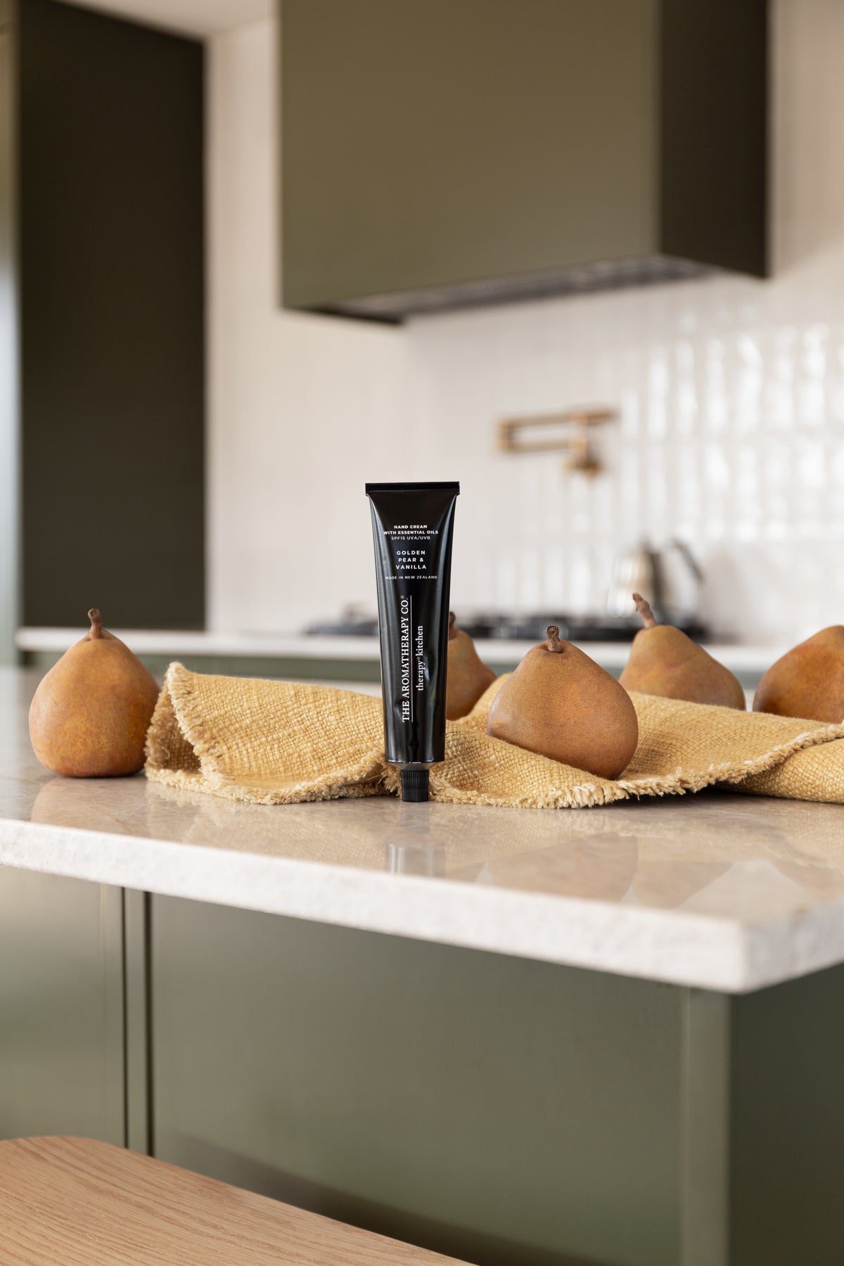 Therapy Kitchen hand cream standing on a stone bench in an olive green kitchen with pears on linen