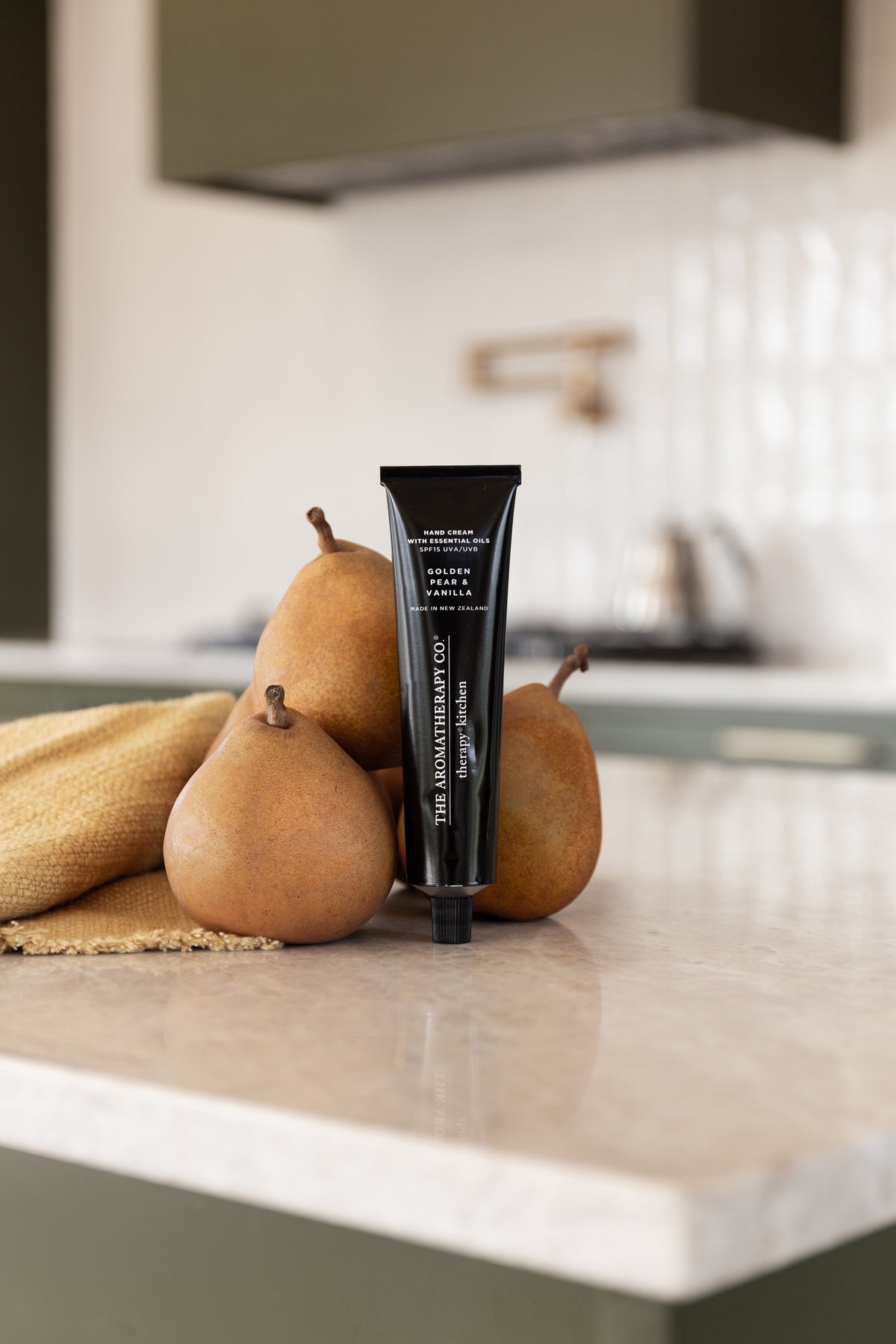 Hand cream tube leaning among three pears on linen at the edge of a stone kitchen bench