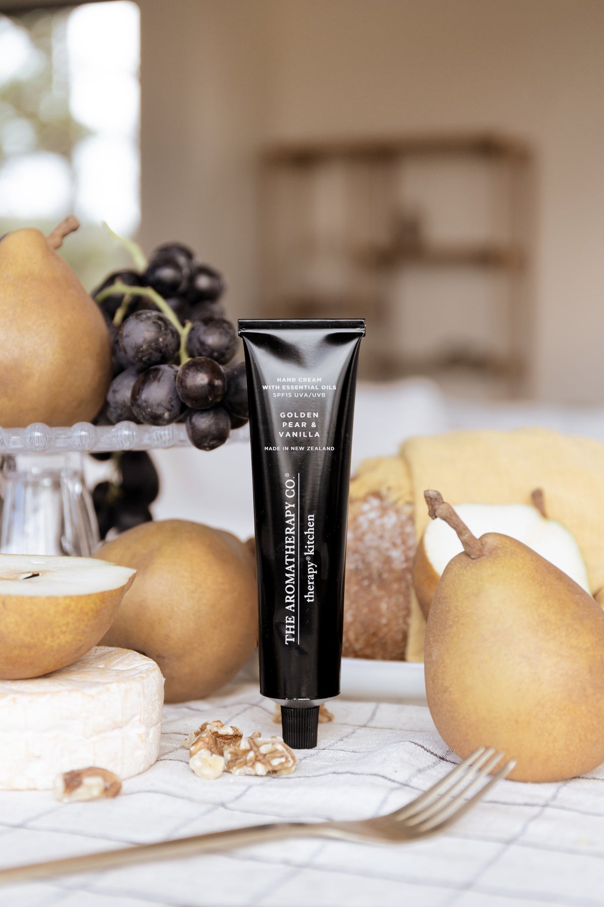 Hand cream tube upright on a table styled with pears, black grapes on a glass stand and bread