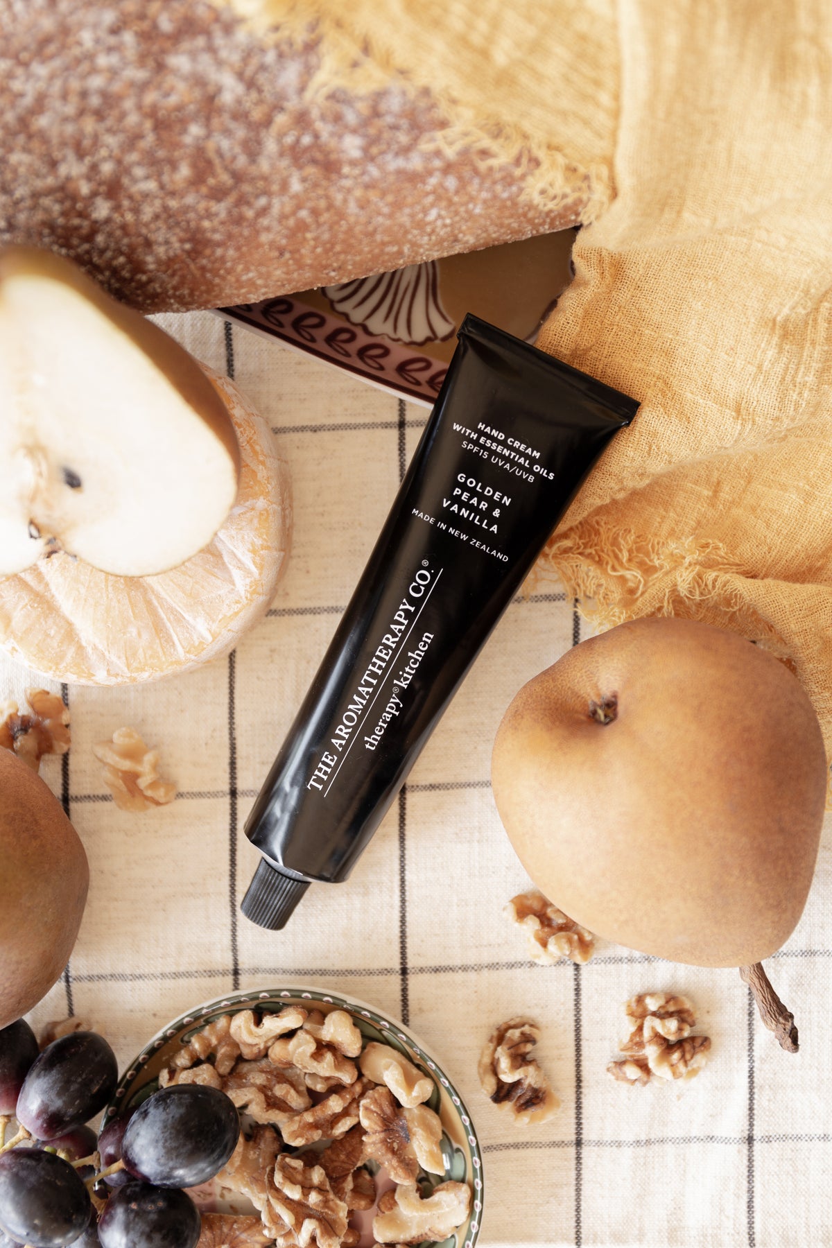 Portrait flatlay of the hand cream tube between a halved pear, a rustic loaf and a bowl of walnuts