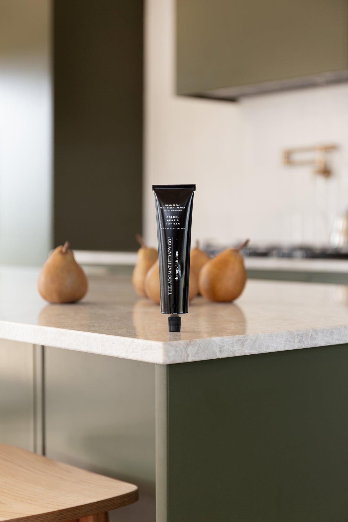 Hand cream tube upright on a marble bench corner with pears behind in an olive green kitchen