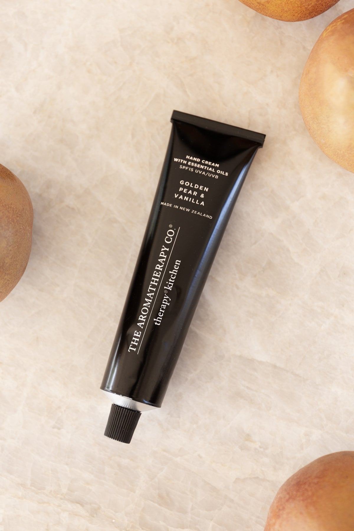 Hand cream tube lying diagonally on pale travertine with whole pears at the edges