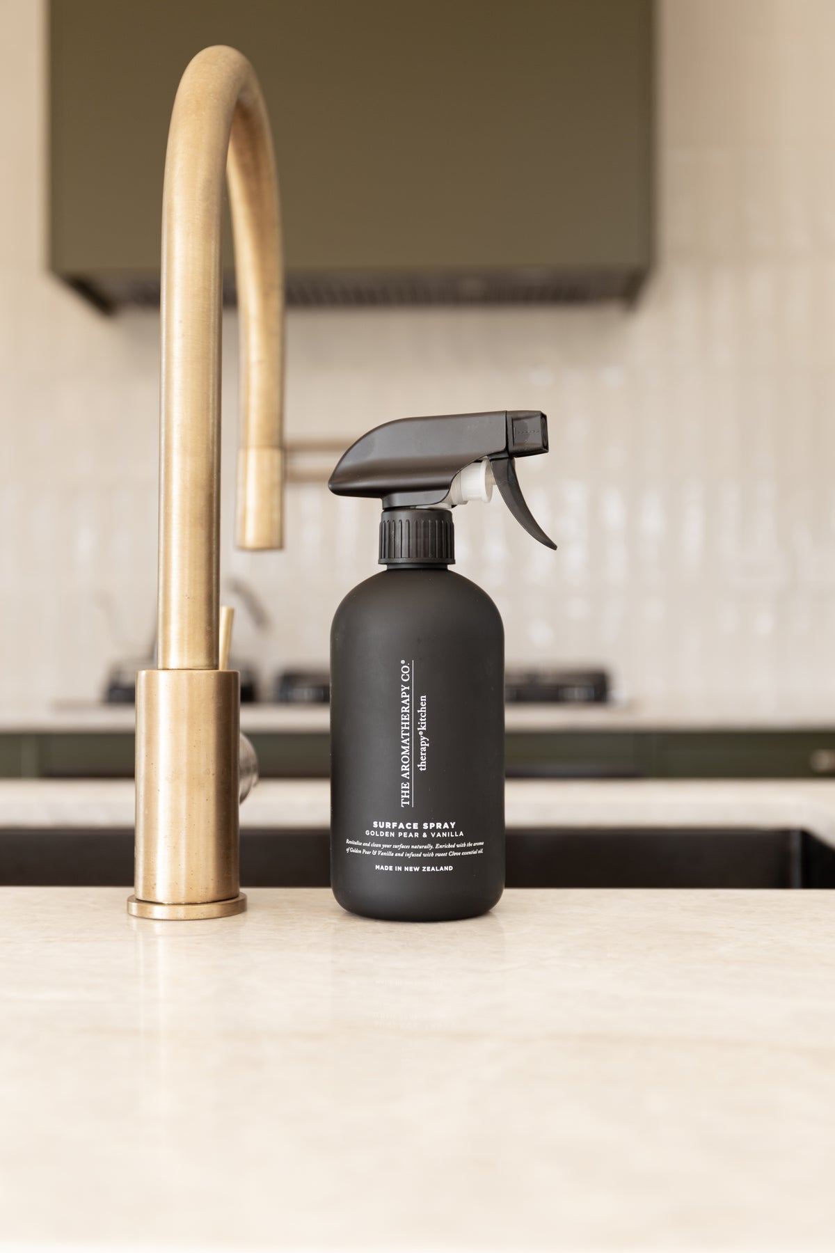 Black surface spray beside a brass gooseneck tap on a stone benchtop with white tiles behind