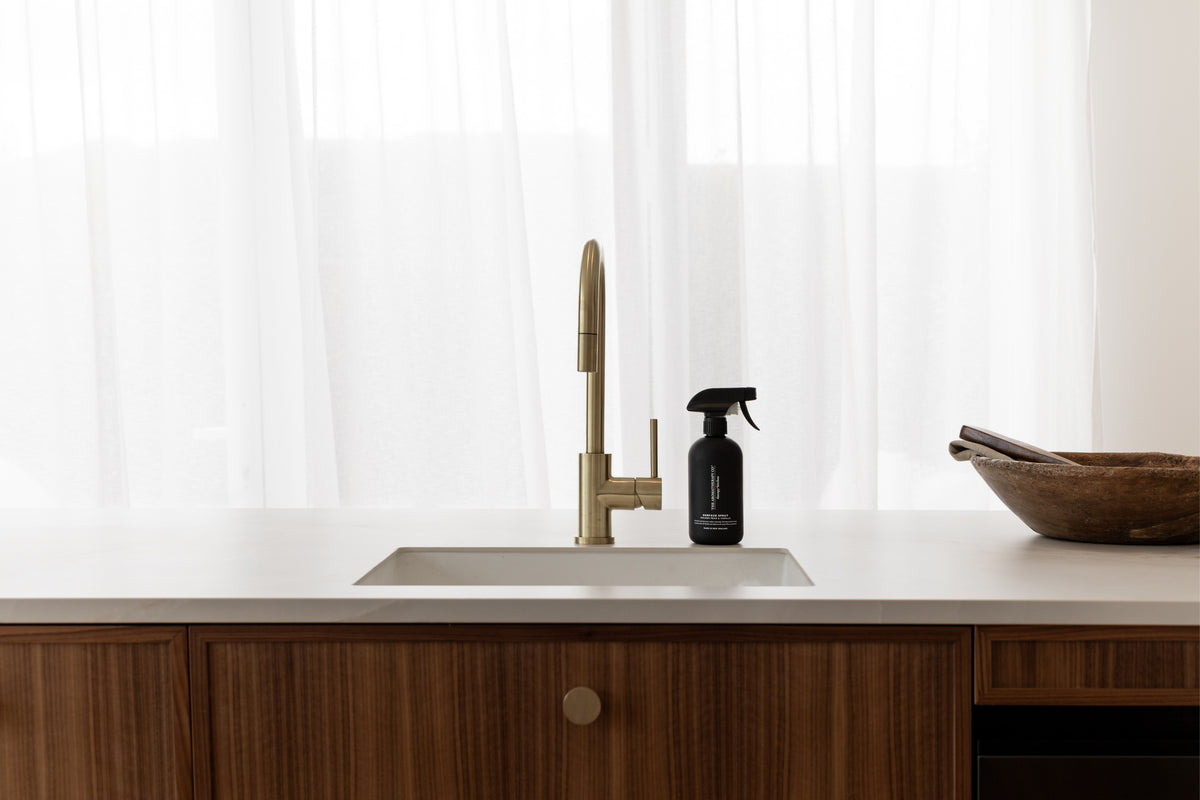 Black surface spray at a white sink with a brass tap, wooden bowl and sheer curtain behind
