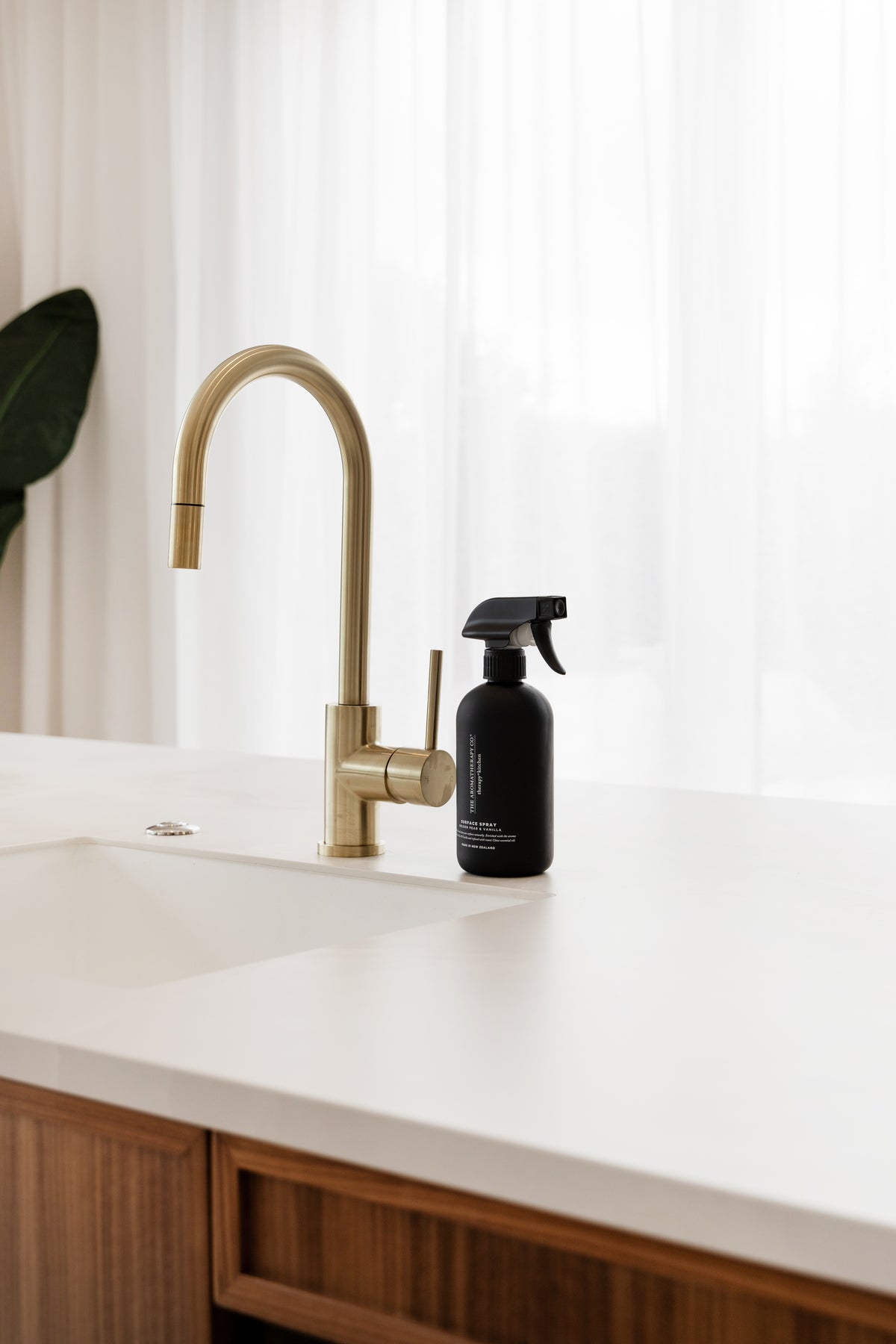 Black surface spray beside a brass tap on a white benchtop with a plant leaf at the edge
