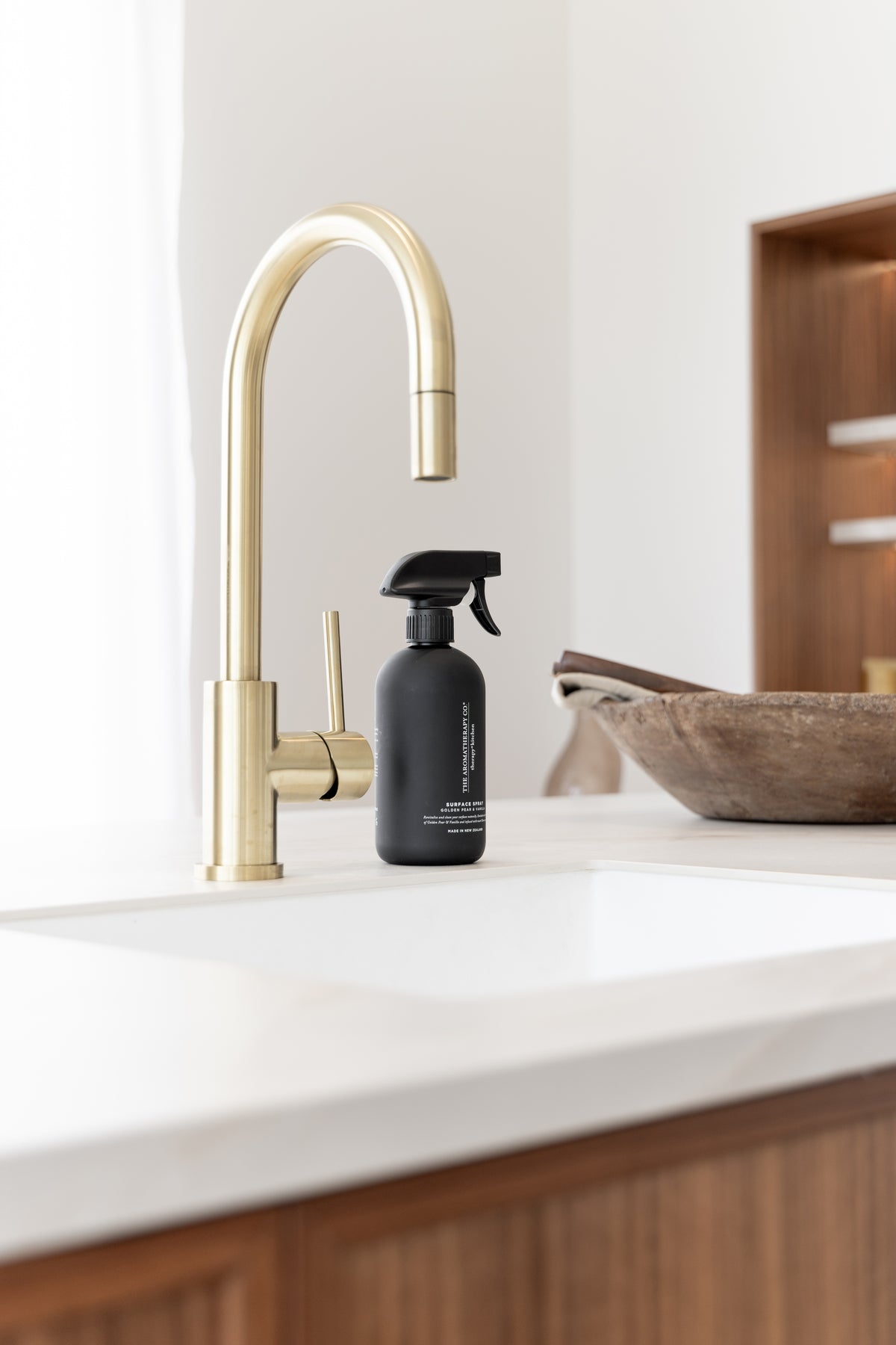 Black surface spray by a brass tap with a carved wooden bowl and timber shelving behind