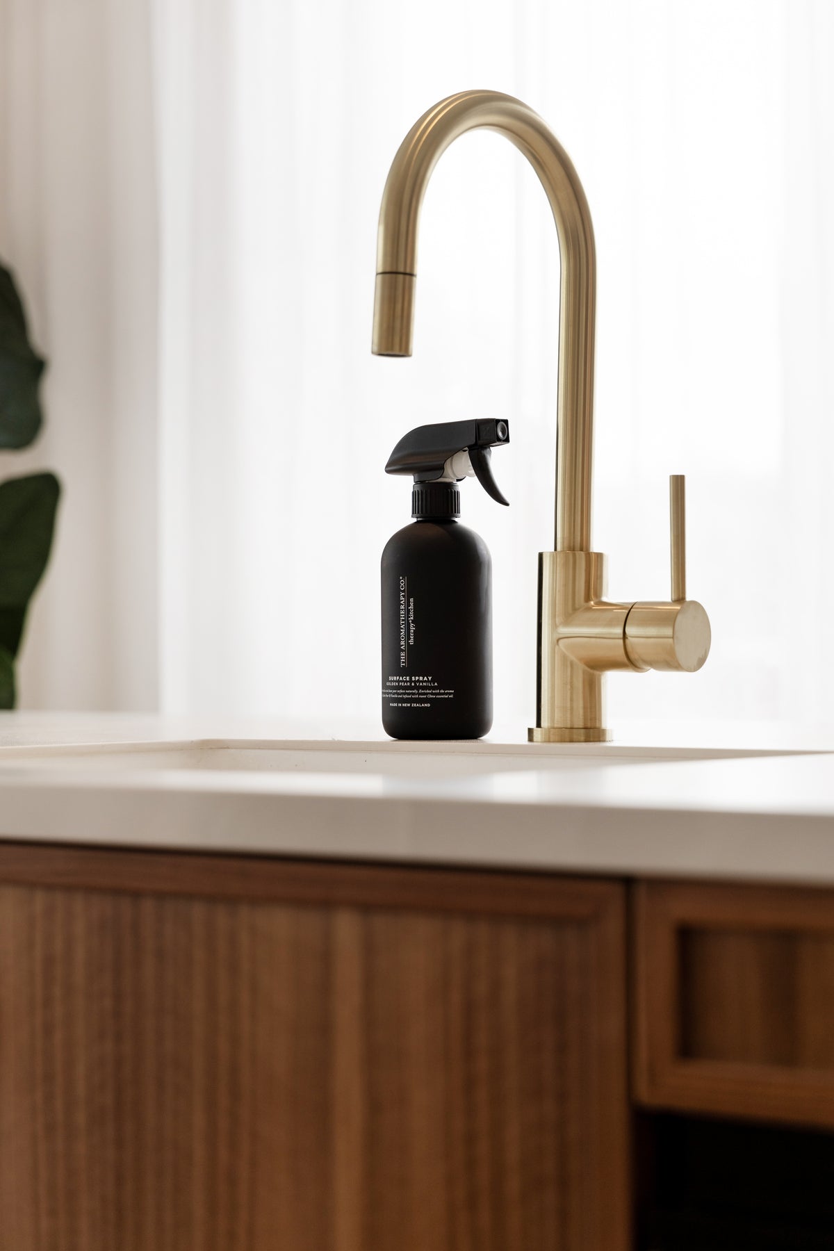 Black surface spray beside a brass tap on a white benchtop with sheer curtains behind