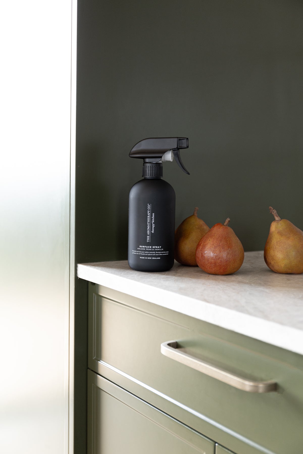Black surface spray on a stone benchtop beside two ripe pears, with olive cabinetry behind