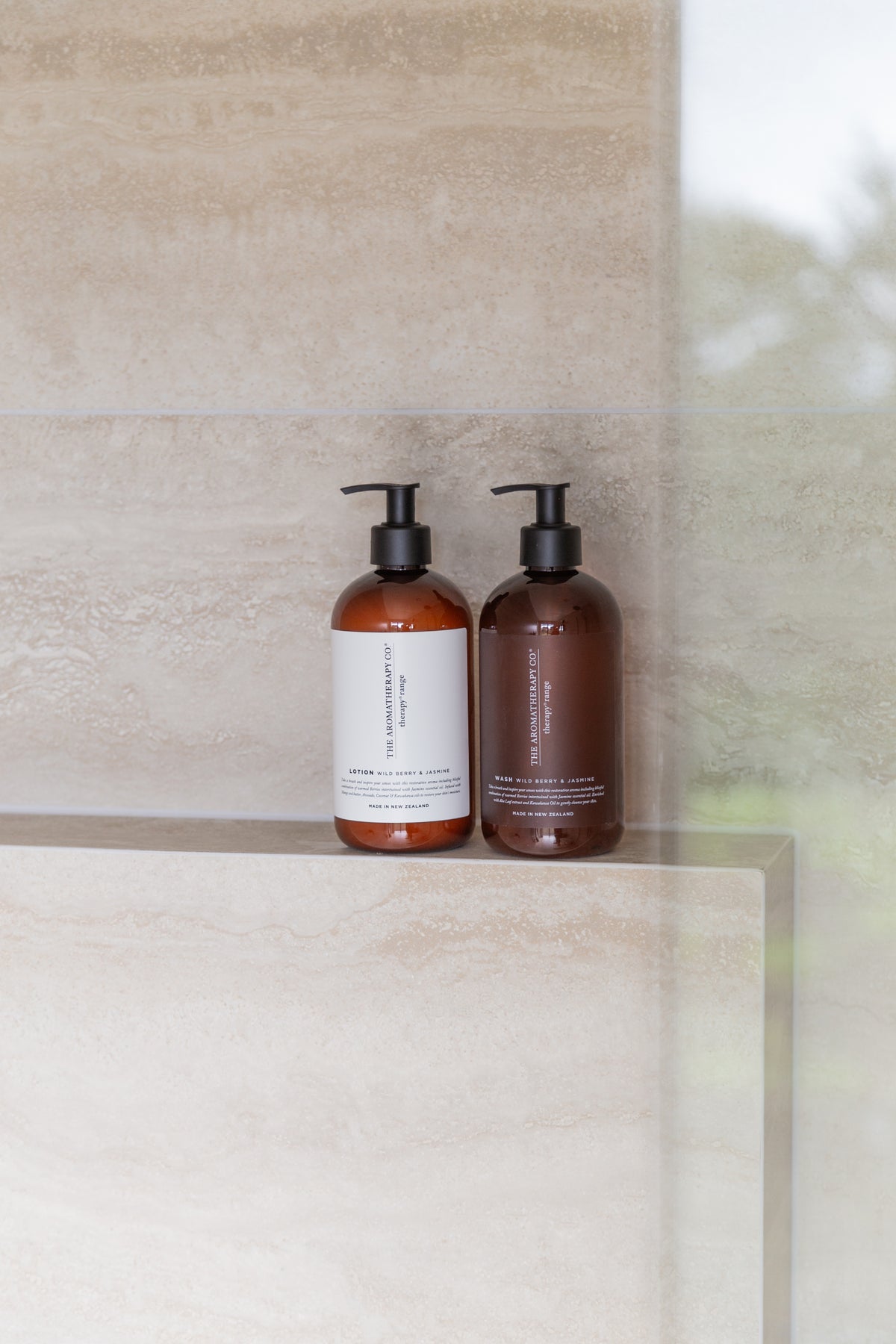 A pair of amber pump bottles on a tiled ledge, seen past the glass panel of a stone tiled shower