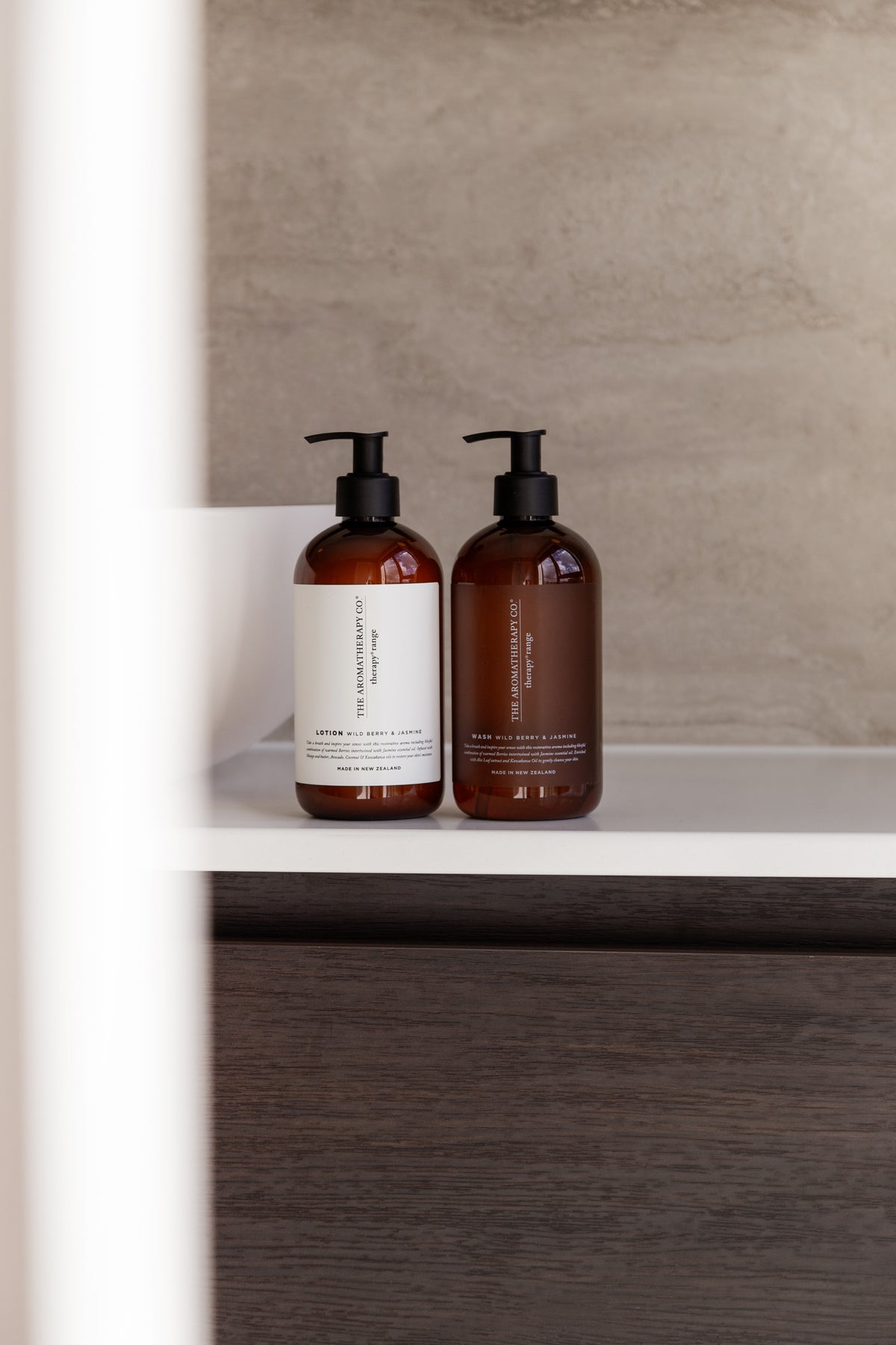 Two amber pump bottles on a white shelf in a stone tiled bathroom, dark timber cabinetry below