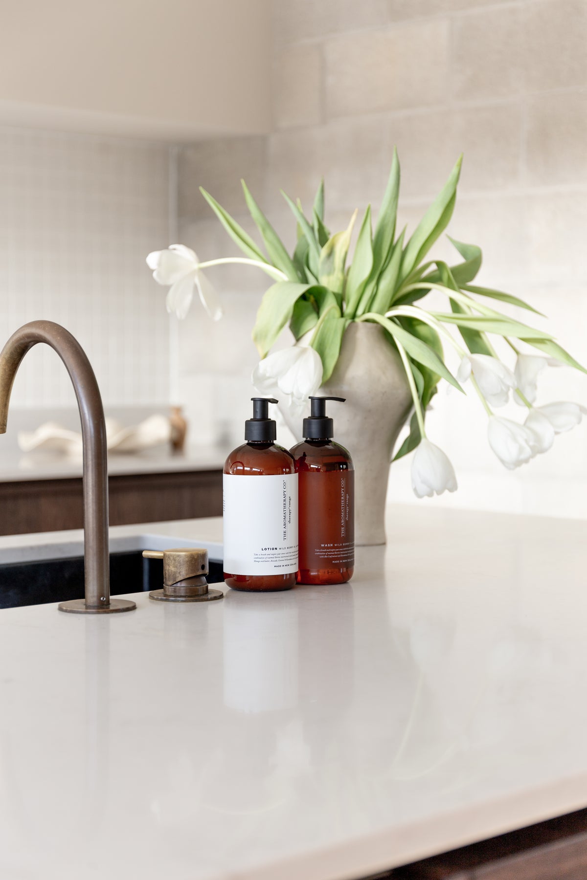 Two amber pump bottles on a white benchtop with a stone vase of white tulips behind them