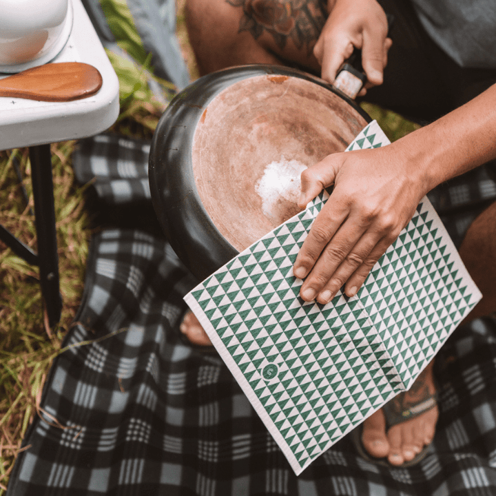 A man is using Good Change's ECO CLOTH - LARGE (2-PACK) to prepare food in a pot on a blanket.