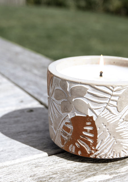 Close view of the lit candle's embossed monstera leaves on a sunlit weathered timber table