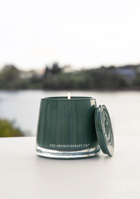 Lit green glass candle with its lid on a white table in front of a blurred lake