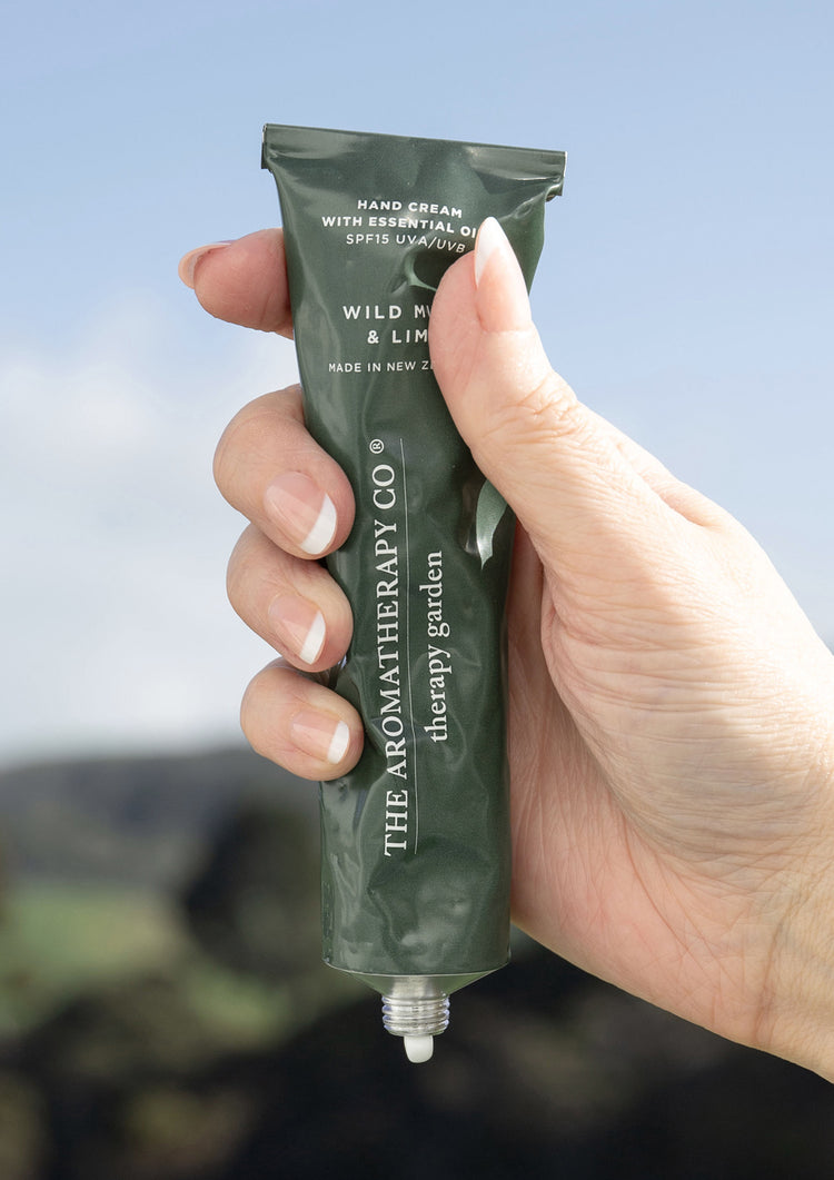 Hand holding the green Therapy Garden hand cream tube up against a blue sky and green hills