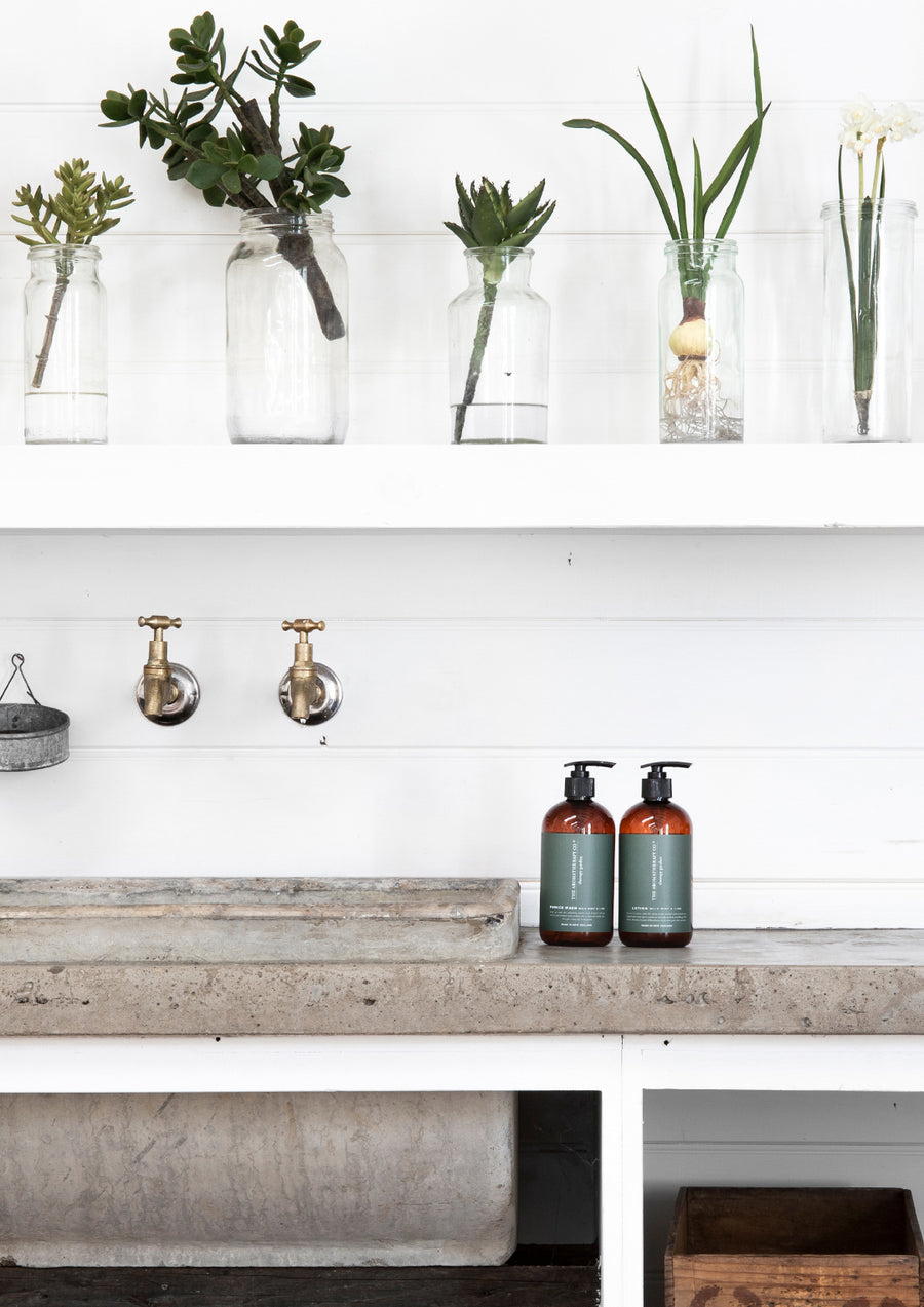 Two amber Therapy Garden bottles on a concrete potting bench below shelves of glass jars with cuttings