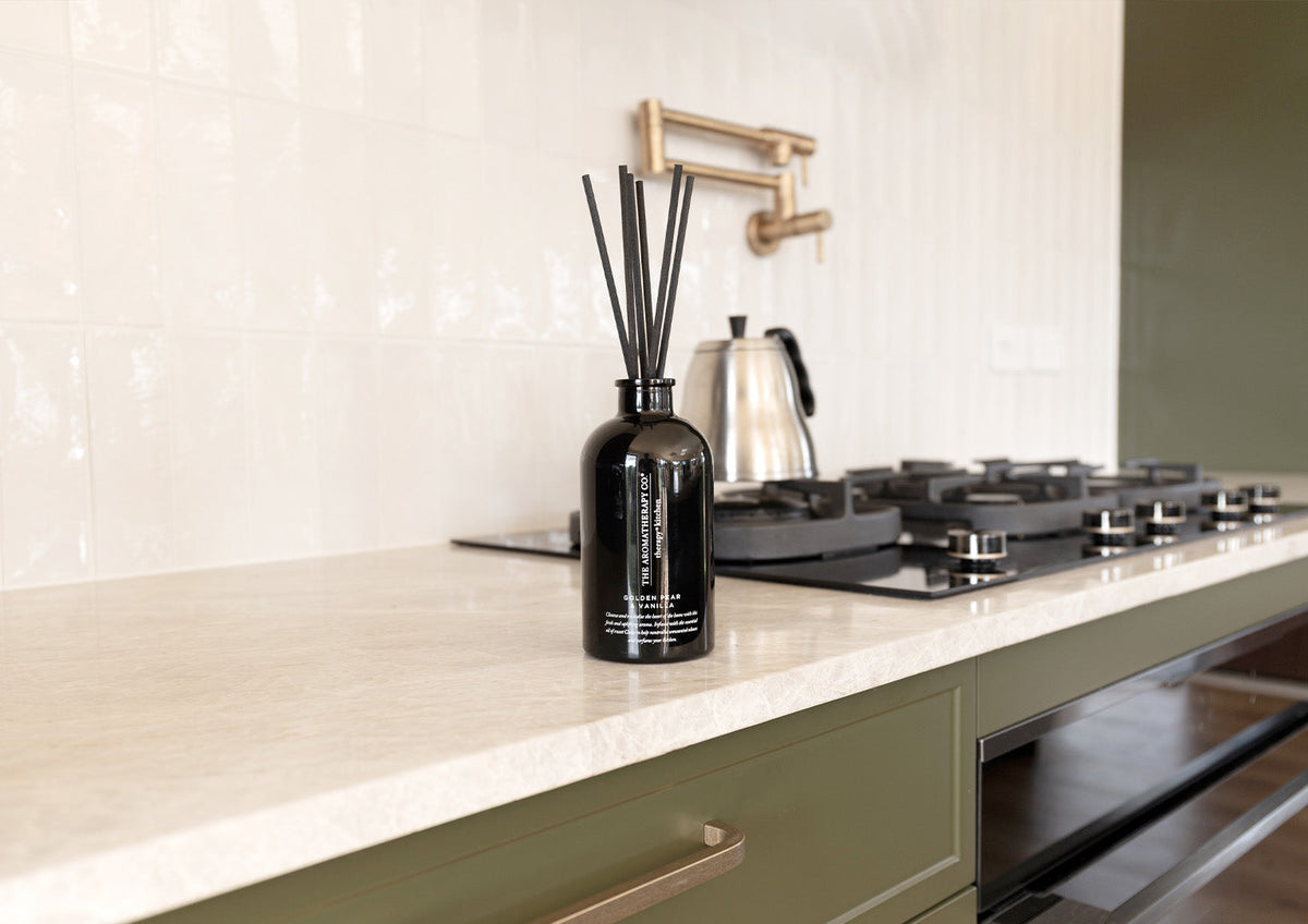 Black reed diffuser beside a gas cooktop on a stone bench in an olive green kitchen, landscape view