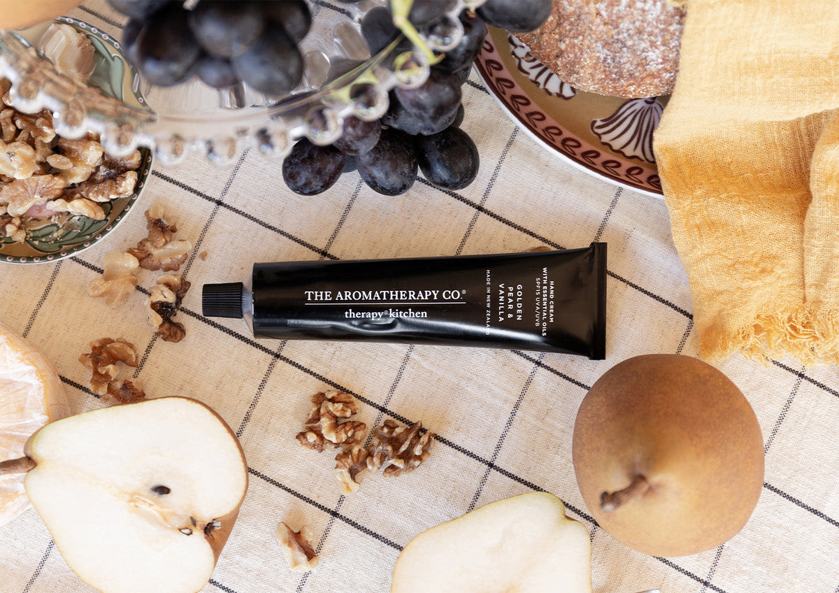 Overhead flatlay of the hand cream tube lying across a check cloth with grapes, walnuts and a halved pear