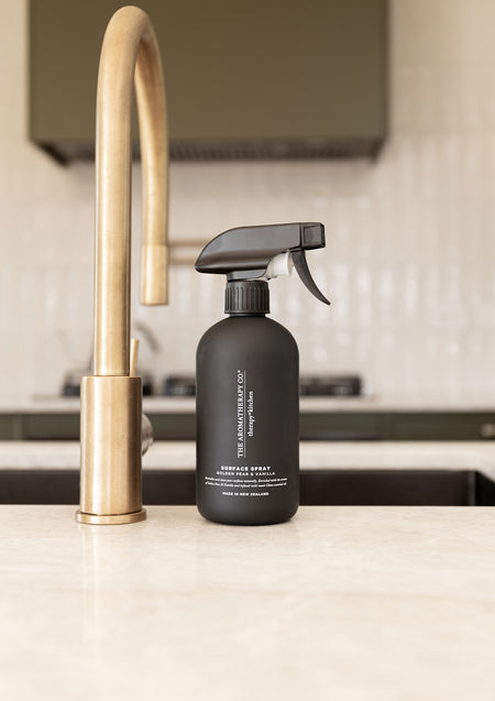 Black surface spray bottle beside a brushed brass gooseneck tap on a pale stone kitchen benchtop