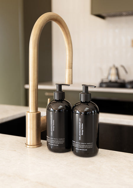 Two black Therapy Kitchen pump bottles beside a brushed brass tap on a pale kitchen benchtop