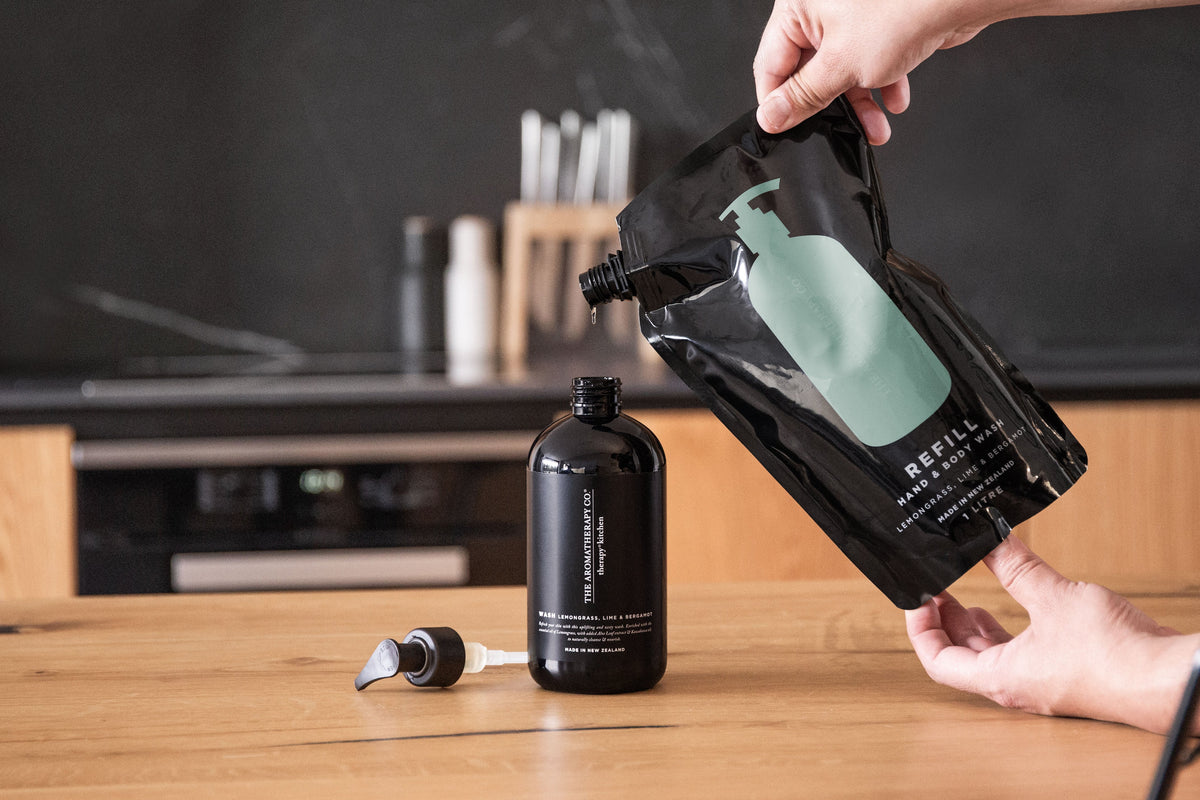 Hands pouring a black refill pouch into a Therapy Kitchen bottle on a timber bench
