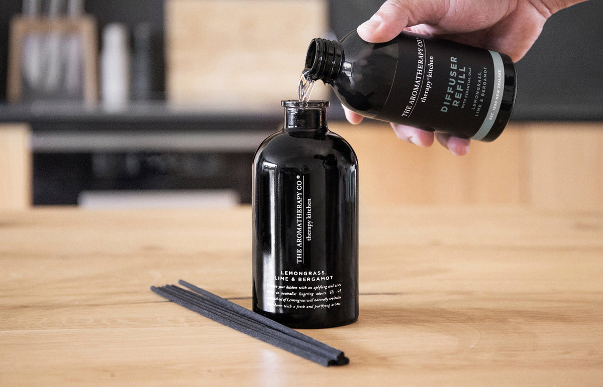 Hand pouring a Therapy Kitchen diffuser refill into a black diffuser bottle on a timber bench