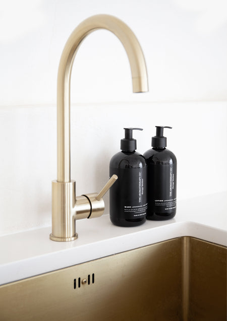Two black Therapy Kitchen pump bottles beside a brass gooseneck tap and brass sink