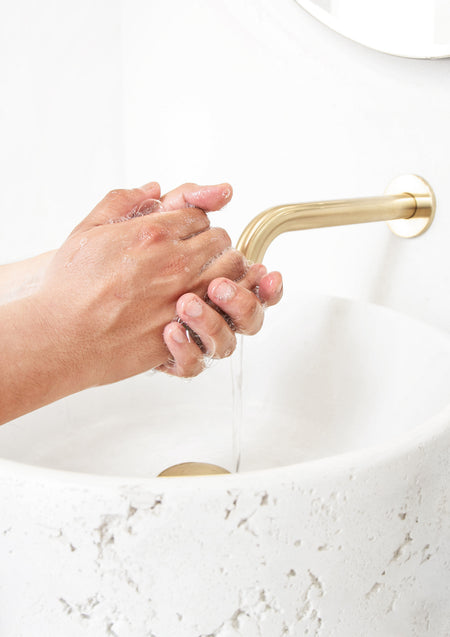 Hands being washed under a brass wall spout over a white stone basin