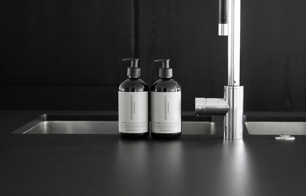 Two Therapy Man pump bottles beside a steel tap at a dark kitchen sink, landscape view