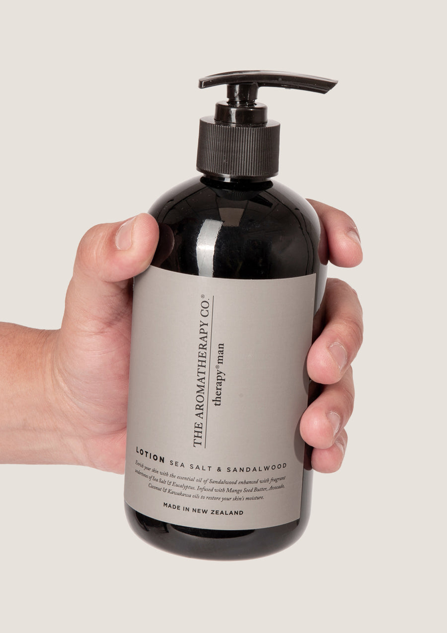Hand holding the Therapy Man Sea Salt & Sandalwood lotion bottle against a pale grey background