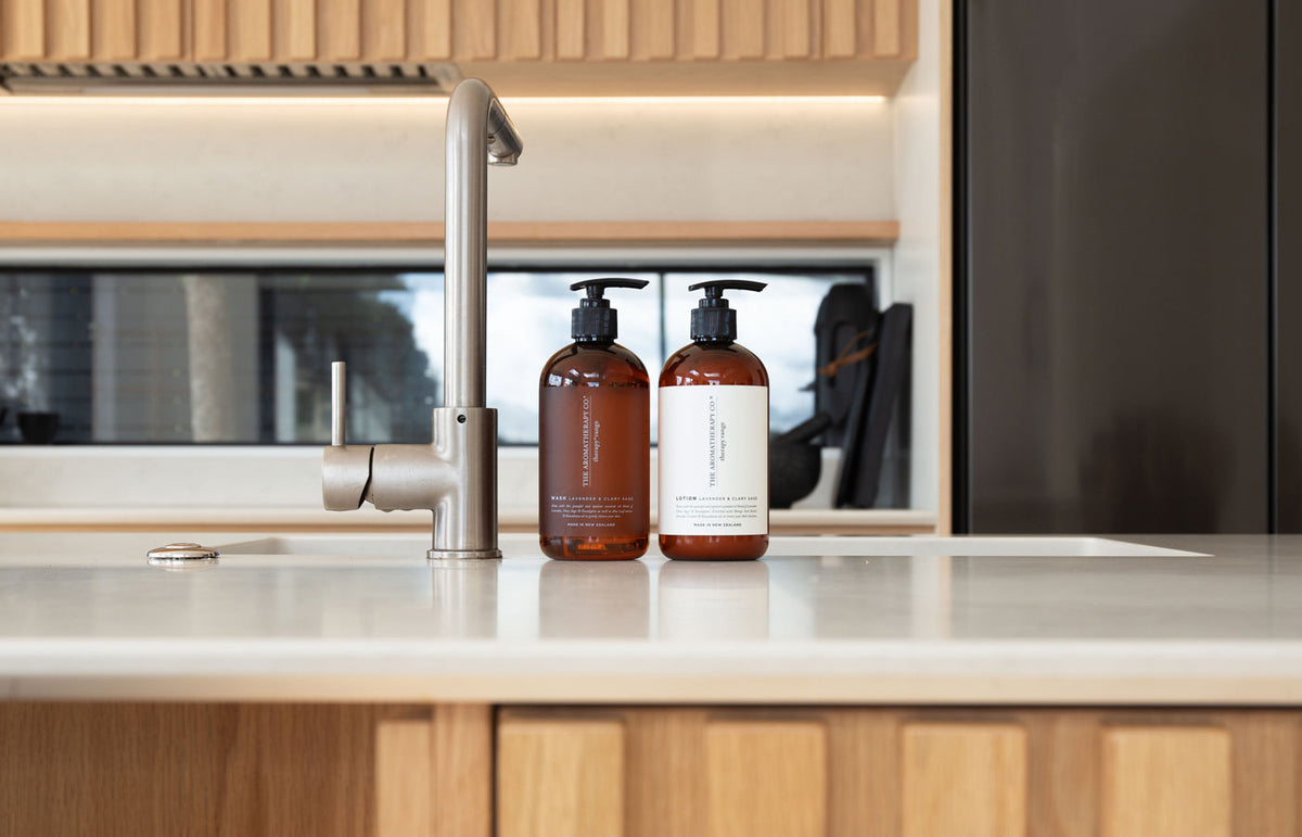 Two amber Therapy pump bottles by a steel tap on a pale bench with a fluted timber cabinet, landscape view