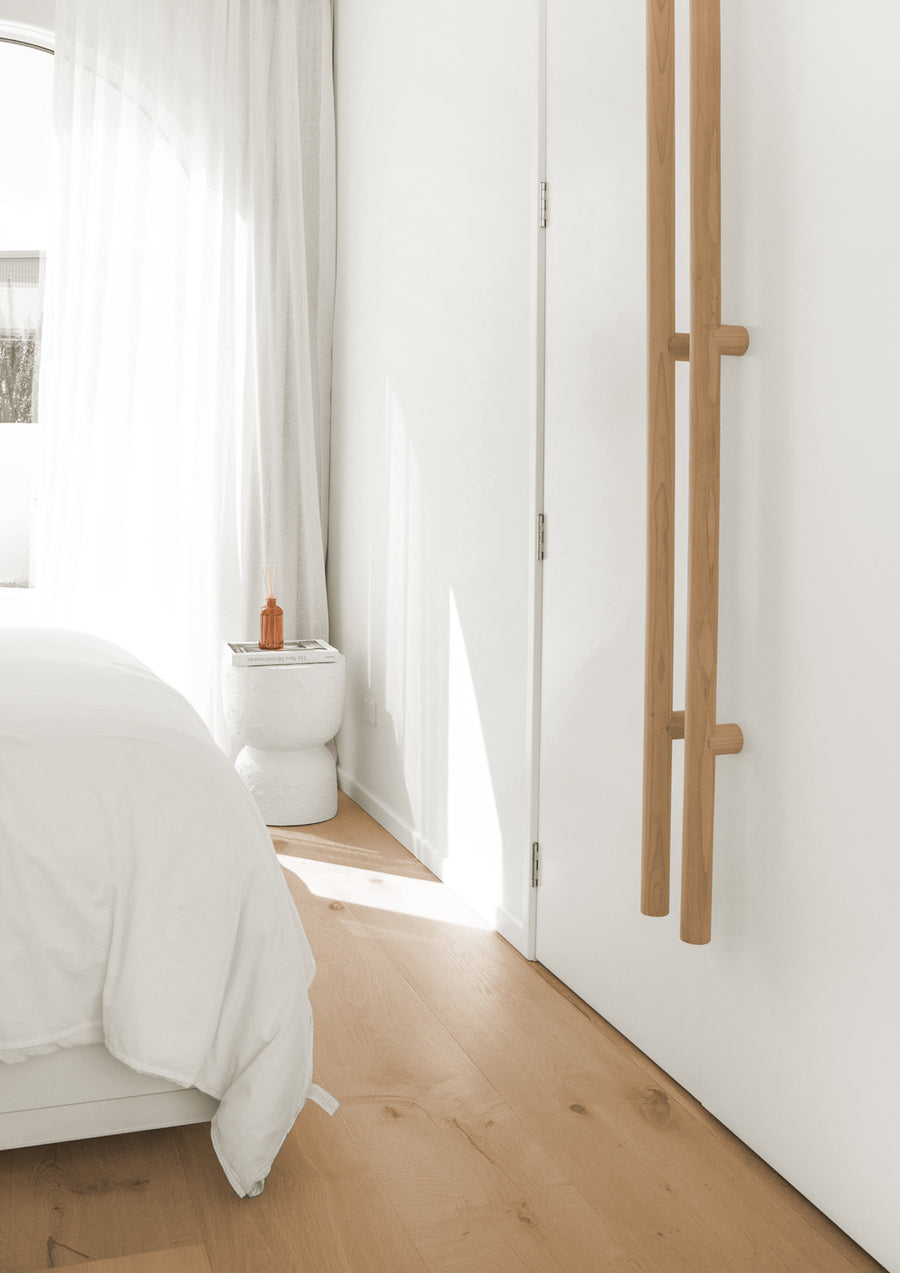 Sunlit bedroom with oak floors, white linen and a timber wall rail, a calm setting for a reed diffuser