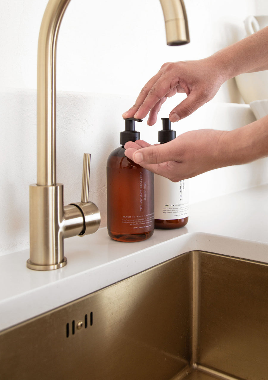 Hands pumping lotion from an amber Therapy bottle beside a brass tap and sink
