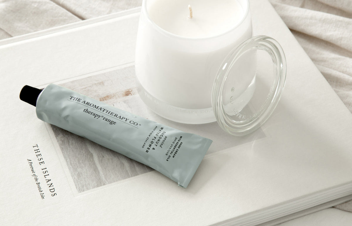 White glass candle with its lid beside a sage hand cream tube on an open book, from above
