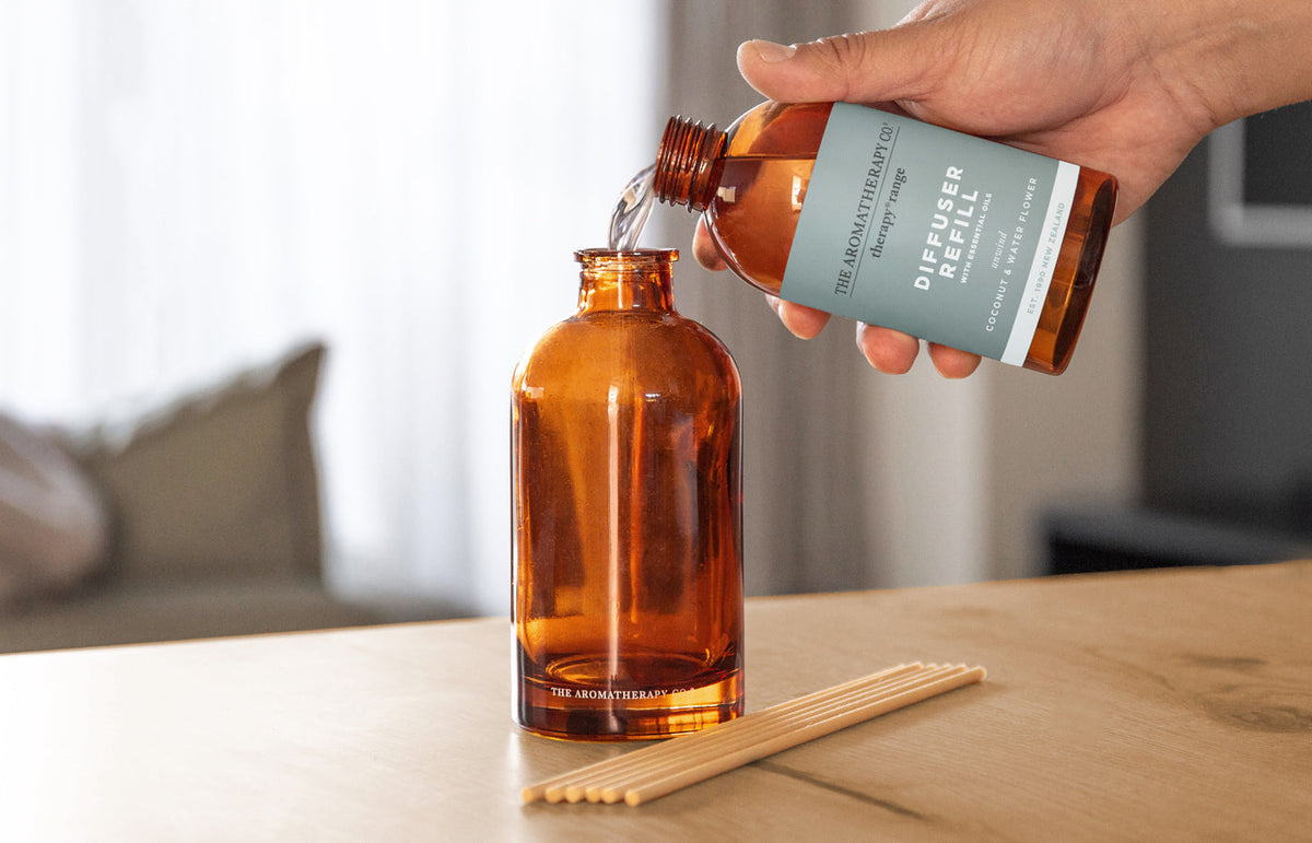 Hand pouring a Therapy Diffuser Refill into the amber diffuser bottle with reeds laid on the table