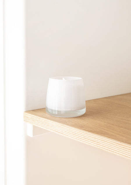 White glass candle on the edge of a pale oak shelf in soft light