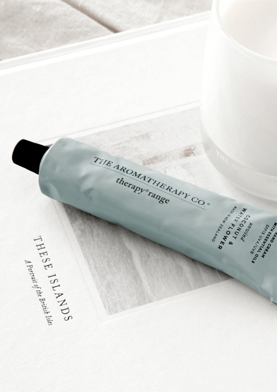 Close up of the sage blue Therapy hand cream tube lying across an open book beside a white cup